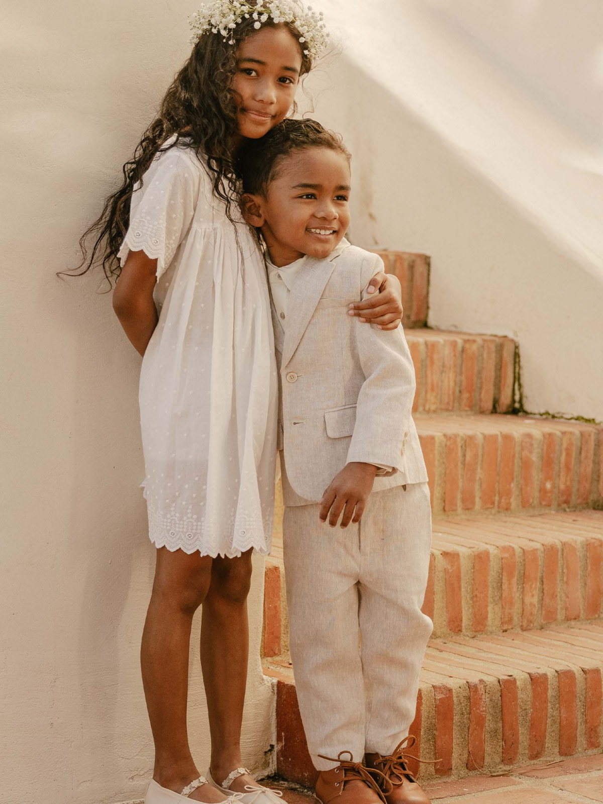 Children smiling together, a girl in a white dress and boy in a beige suit, standing on stairs with a rustic background.
