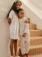 Children smiling together, a girl in a white dress and boy in a beige suit, standing on stairs with a rustic background.