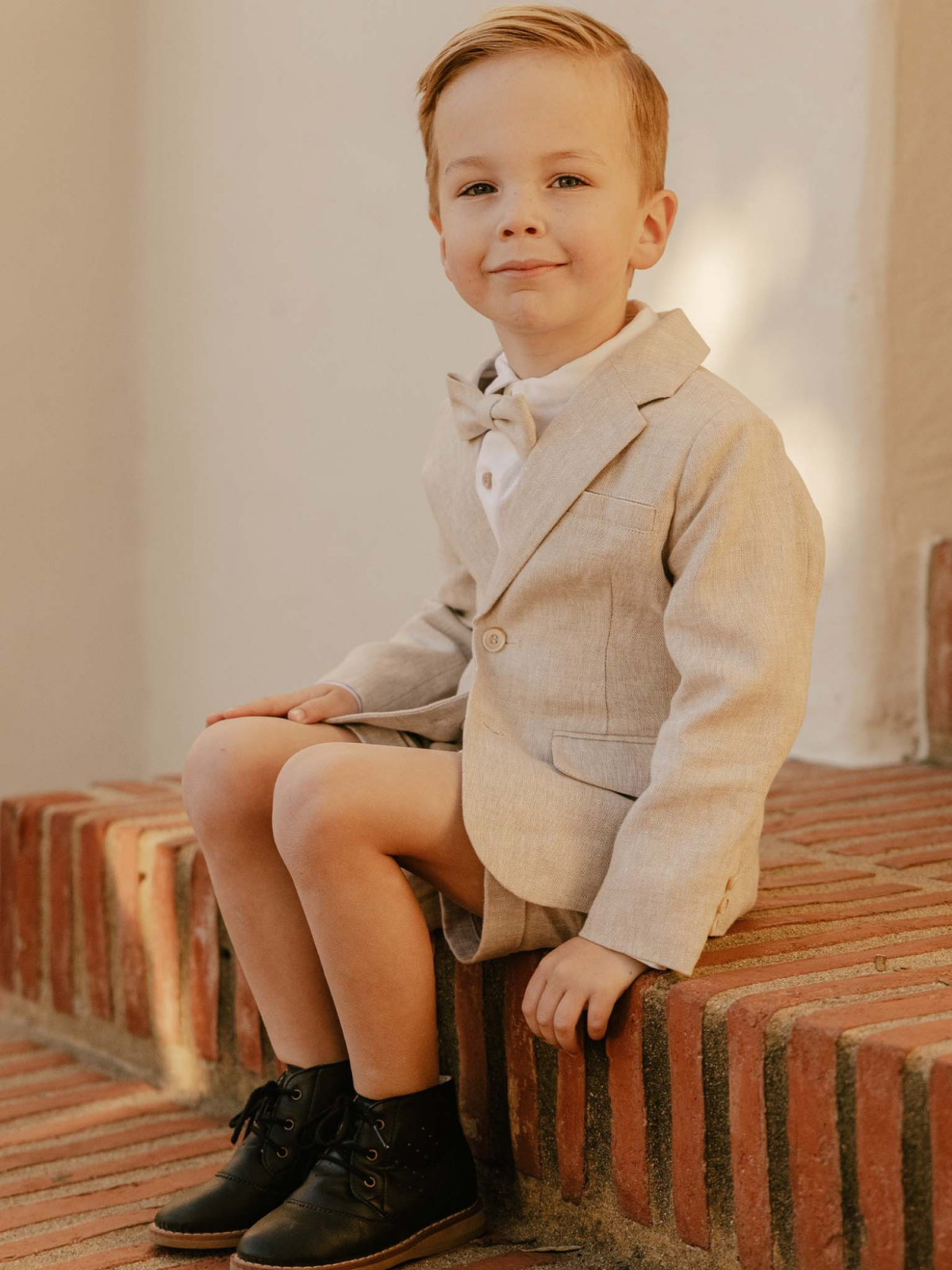 Smiling boy in a light suit sits on brick steps, showcasing a stylish outfit and black boots.