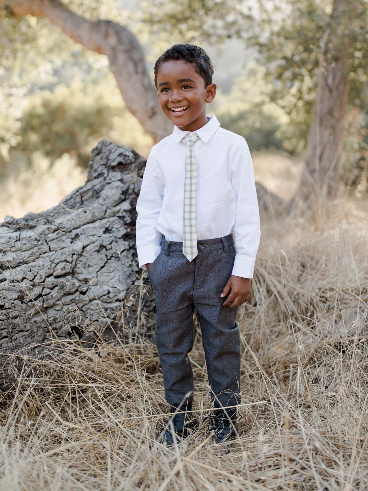 Smiling boy in formal outfit standing in grass near a tree, wearing a white shirt, gray pants, and a patterned tie.