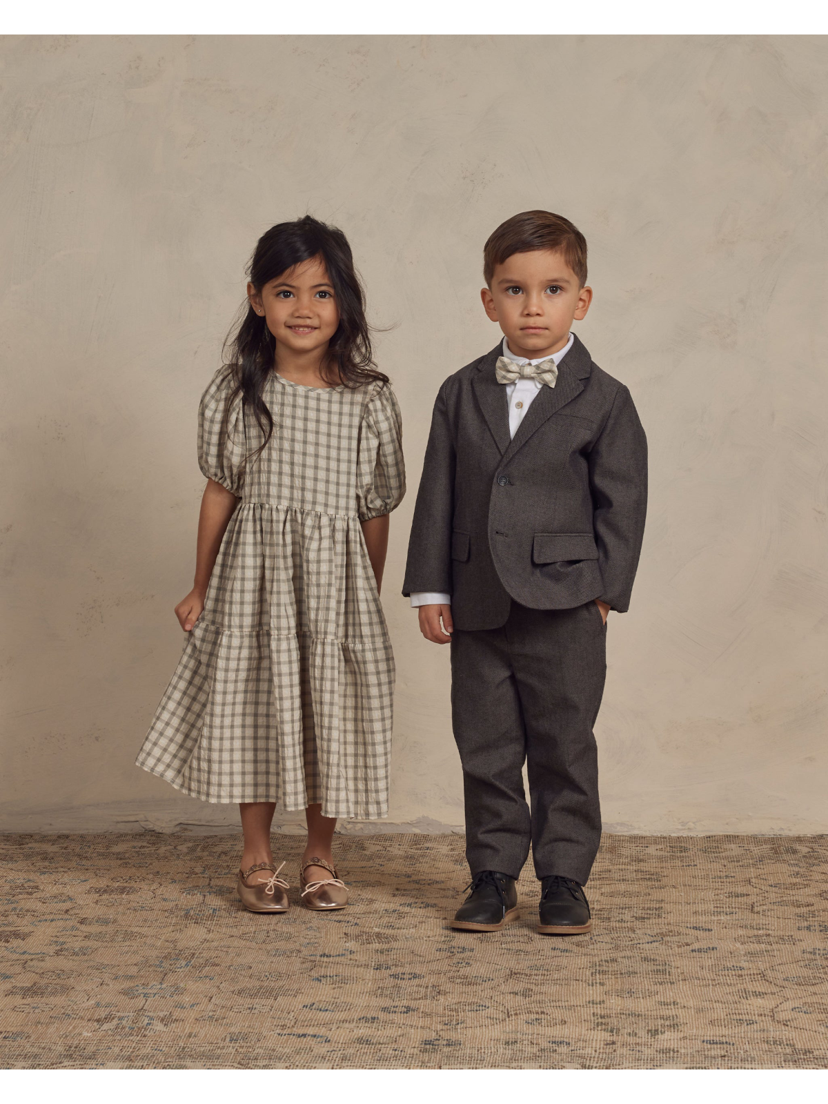 Two children stand side by side: a girl in a plaid dress and a boy in a suit with a bowtie, against a neutral background.