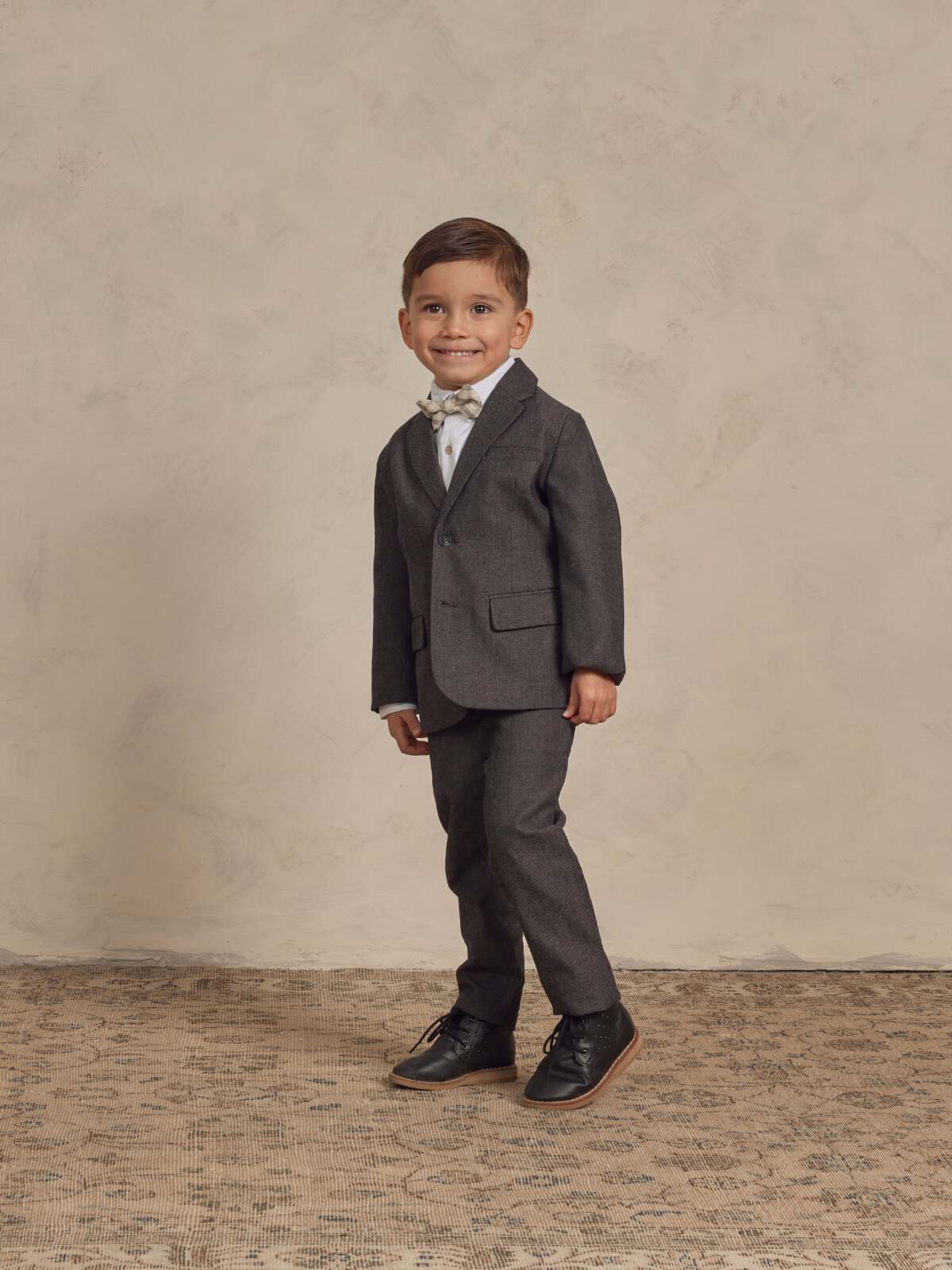 Smiling boy in a gray suit with a bow tie, standing on a textured floor against a neutral background.