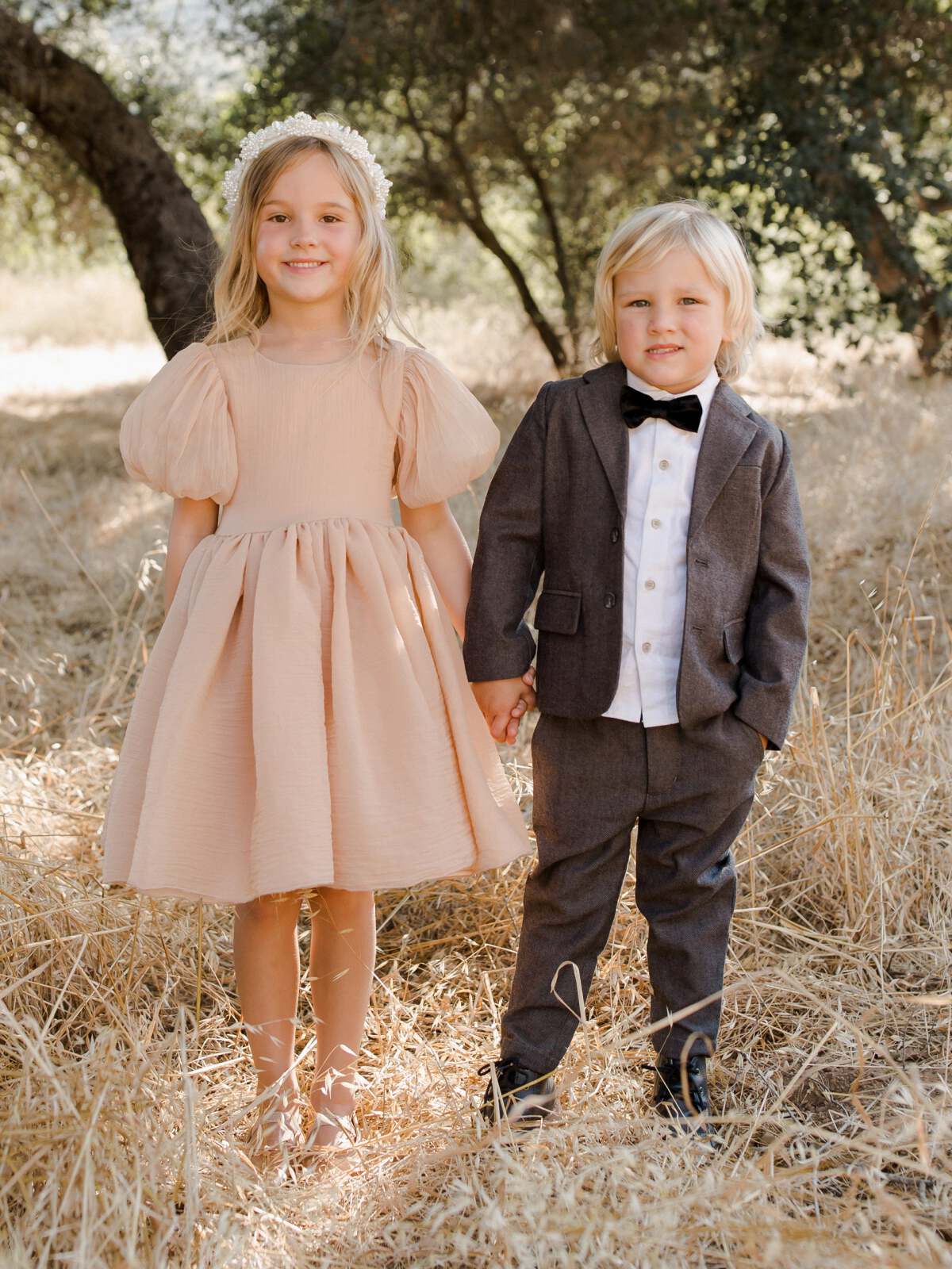Two young children, a girl in a pink dress and a boy in a suit, holding hands in a grassy field.