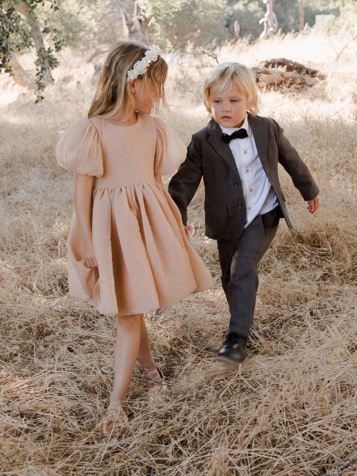 Two children walk together in a field of dry grass, wearing a dress and suit in soft tones.