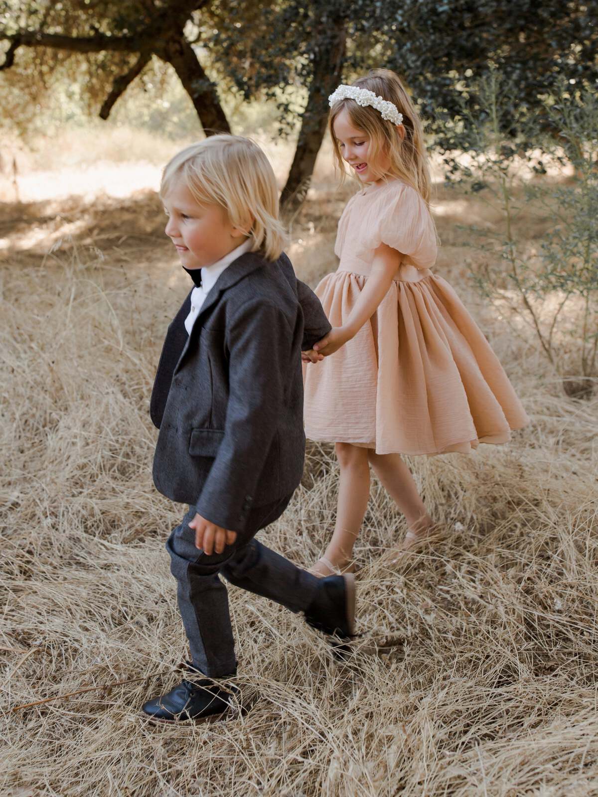 Two young children walk through tall grass; a boy in a suit and a girl in a dress, enjoying a playful moment outdoors.