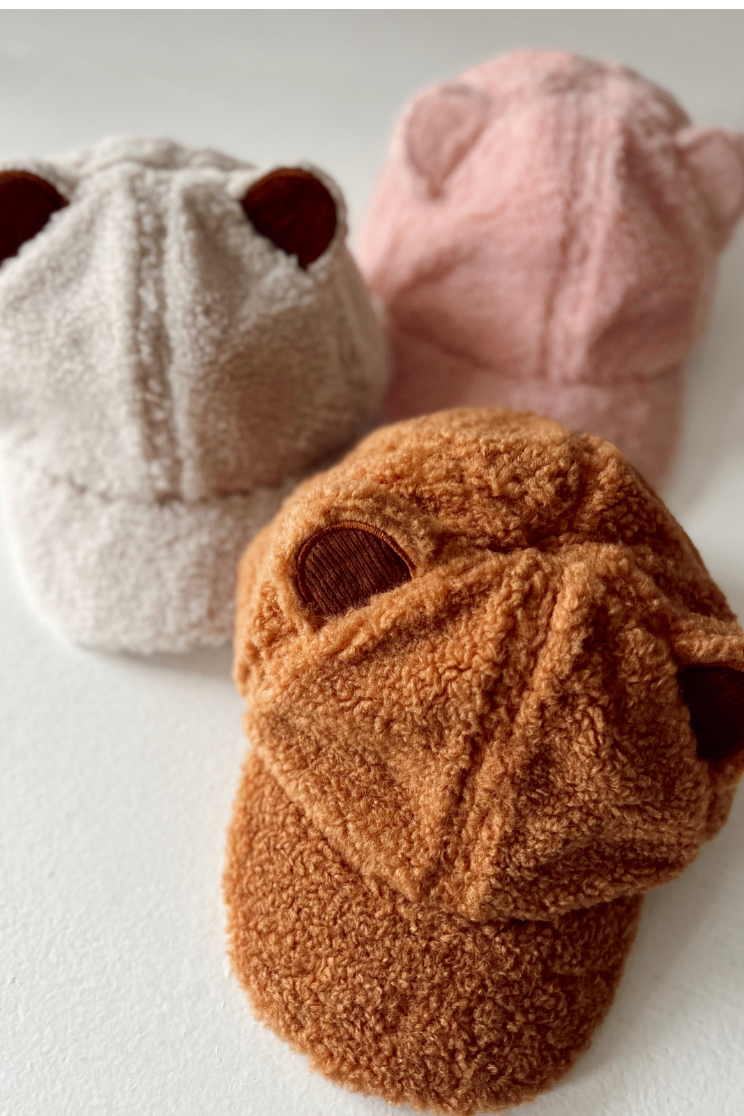 Three cozy hats with ear details in white, pink, and orange, displayed on a white surface.