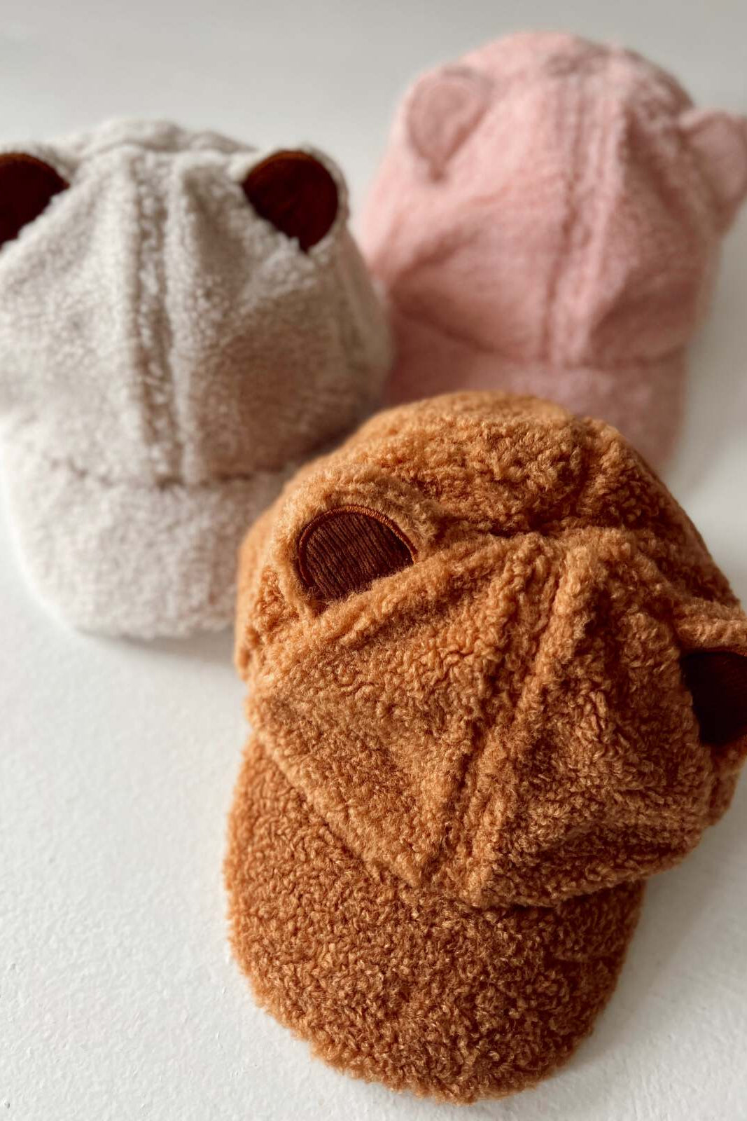 Three fuzzy bear-ear hats in beige, pink, and brown stacked on a white surface.