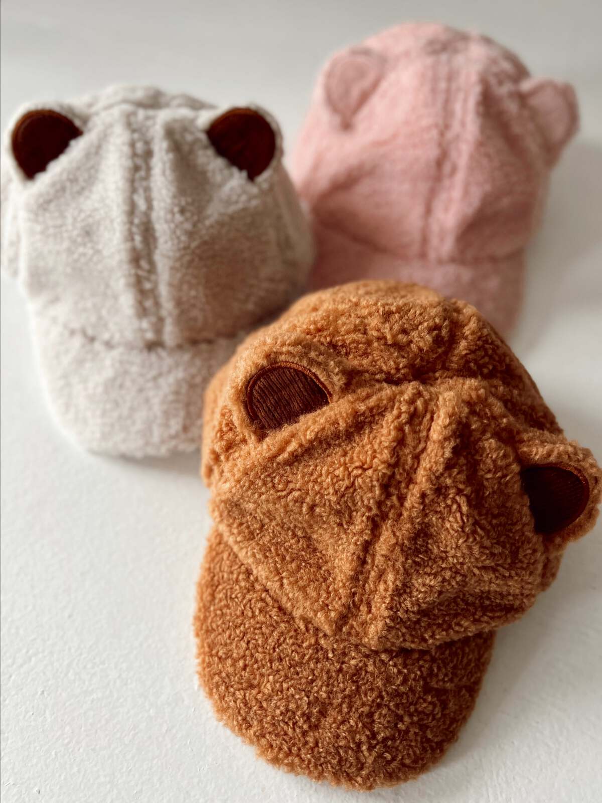 Three cozy bear ear hats in beige, pink, and brown plush fabric on a white surface.