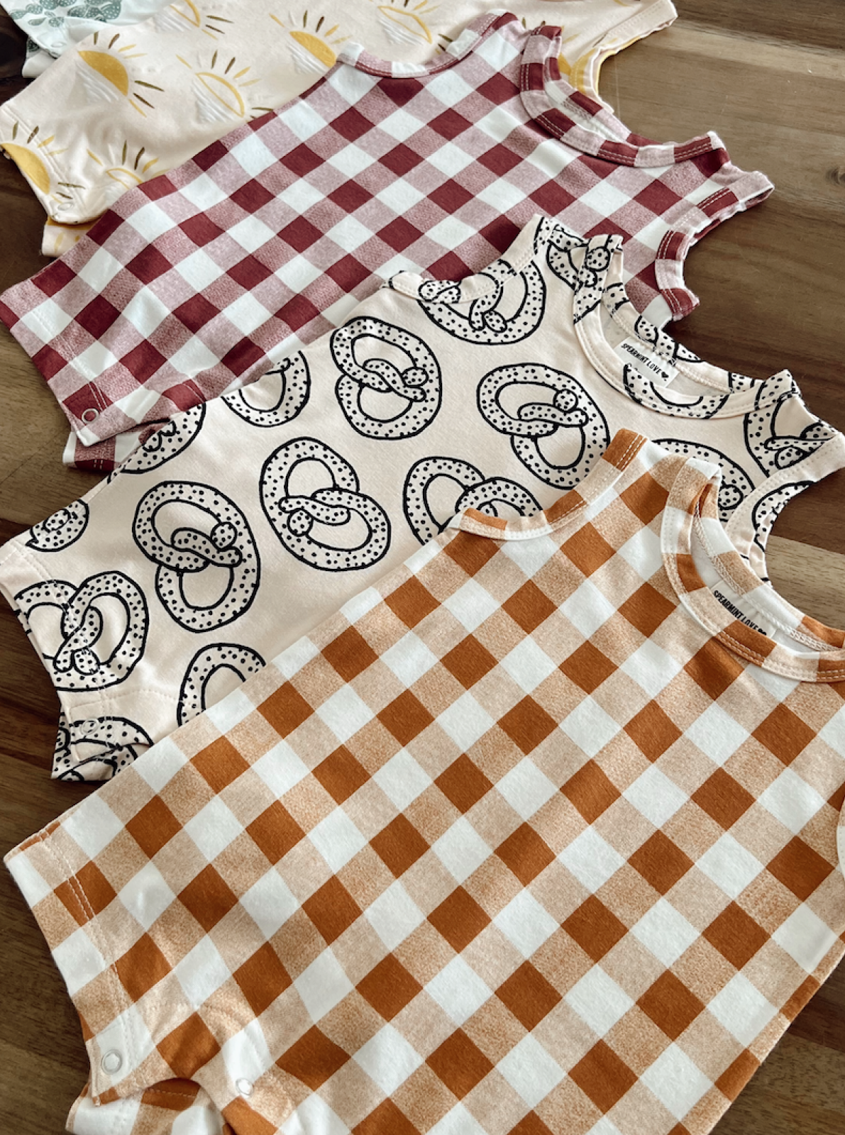 Four patterned sleeveless baby shirts displayed on a wooden surface, featuring checks and pretzel designs.