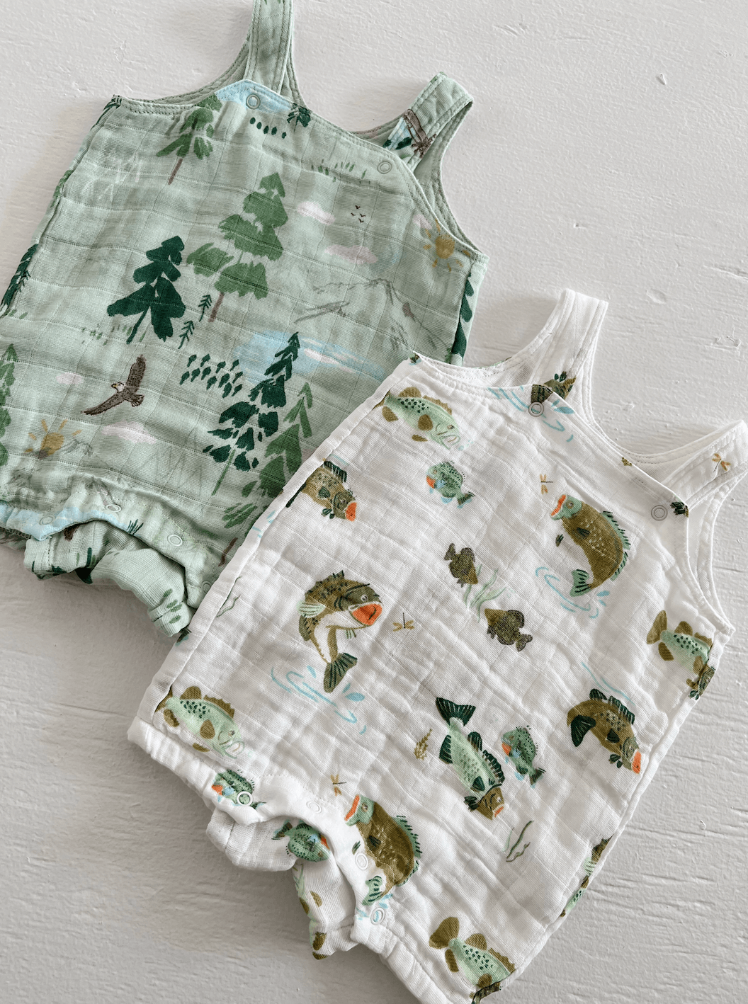 Two colorful baby rompers with nature-themed prints: trees on green and fish on white, displayed on a light background.