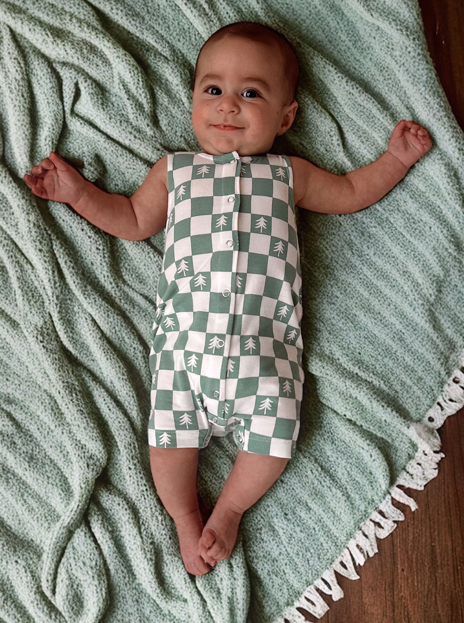 Infant lying on a green blanket, wearing a checkered onesie with tree patterns, smiling at the camera.