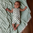 Infant lying on a green blanket, wearing a checkered onesie with tree patterns, smiling at the camera.
