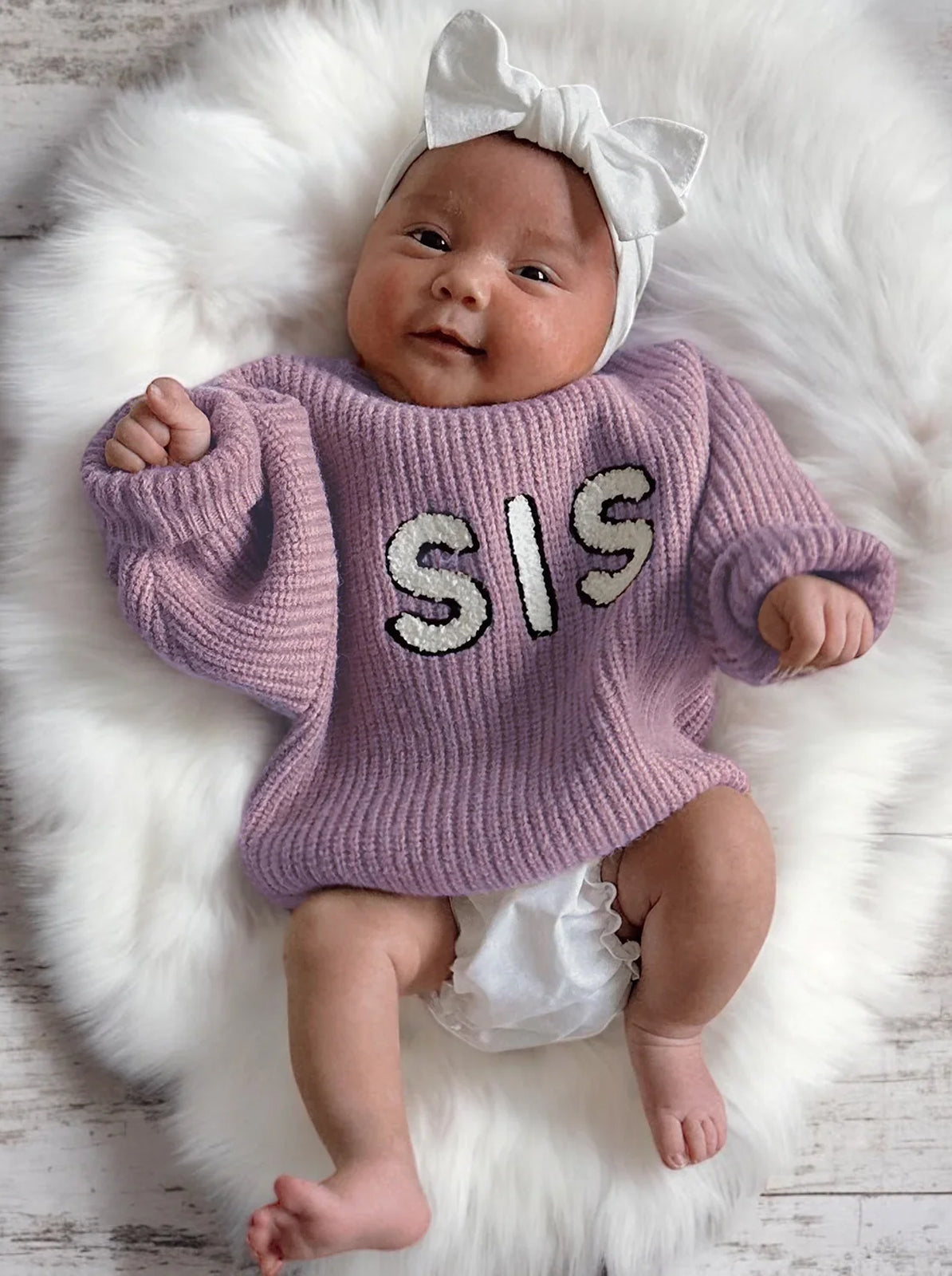 Cute baby in a purple "SIS" sweater with a white bow on a fluffy backdrop, smiling happily.
