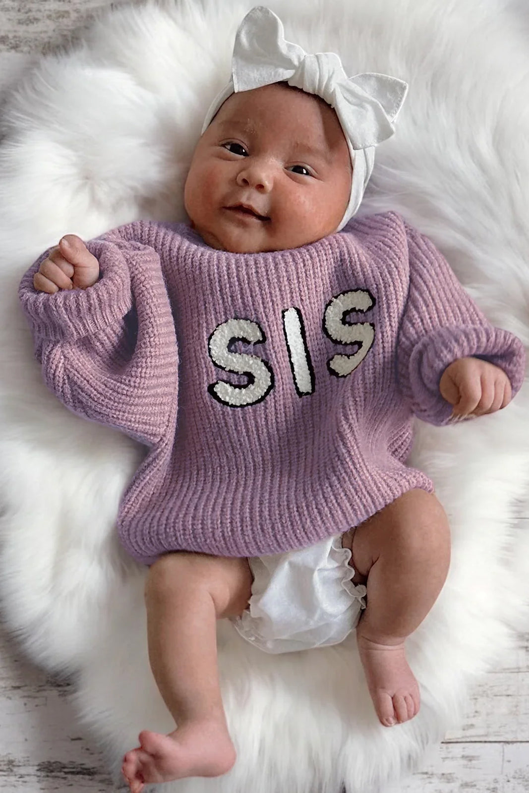 Cute baby in a purple "SIS" sweater with a white bow on a fluffy backdrop, smiling happily.