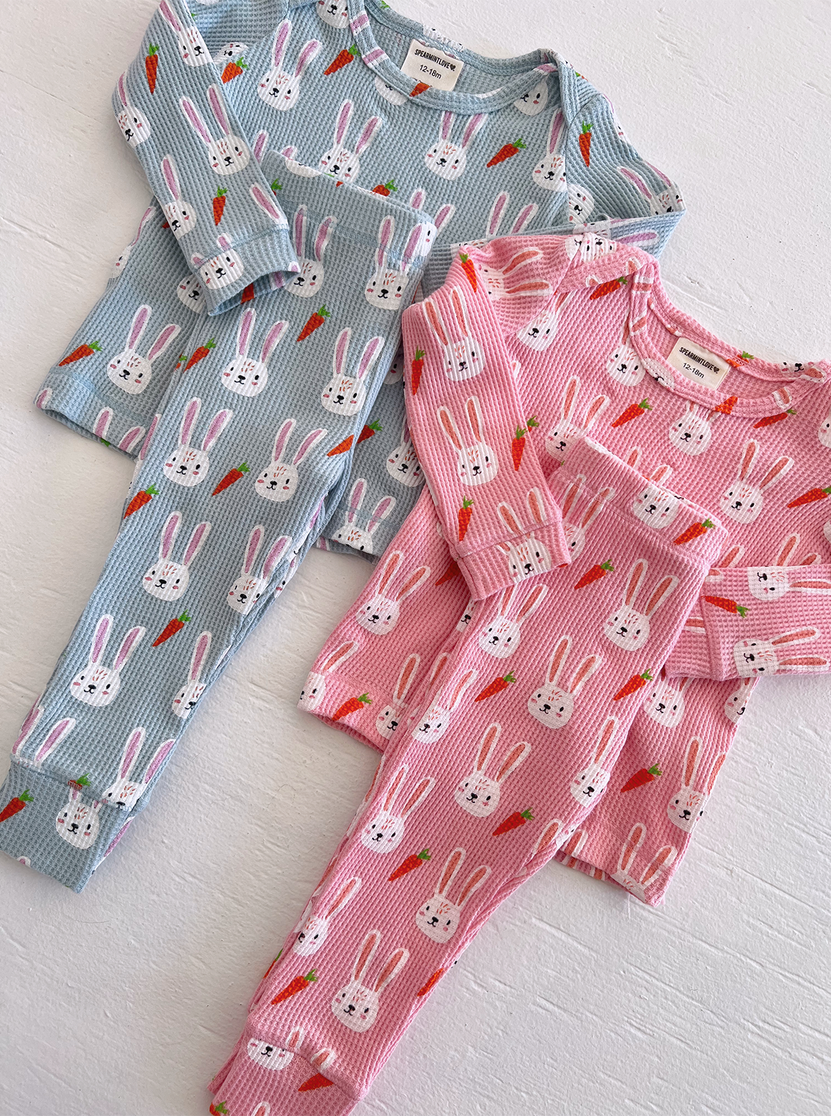 Two sets of children's pajamas featuring bunny and carrot patterns, one in blue and one in pink.