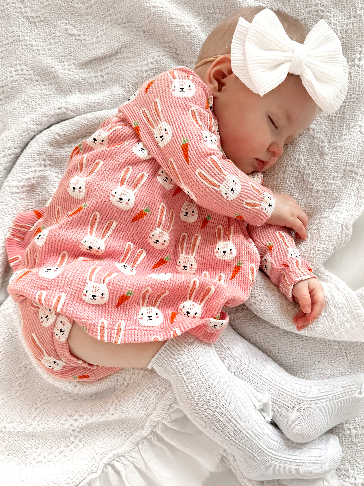 Infant sleeping peacefully on a blanket, wearing a pink bunny-patterned outfit and white bow, with cozy white socks.