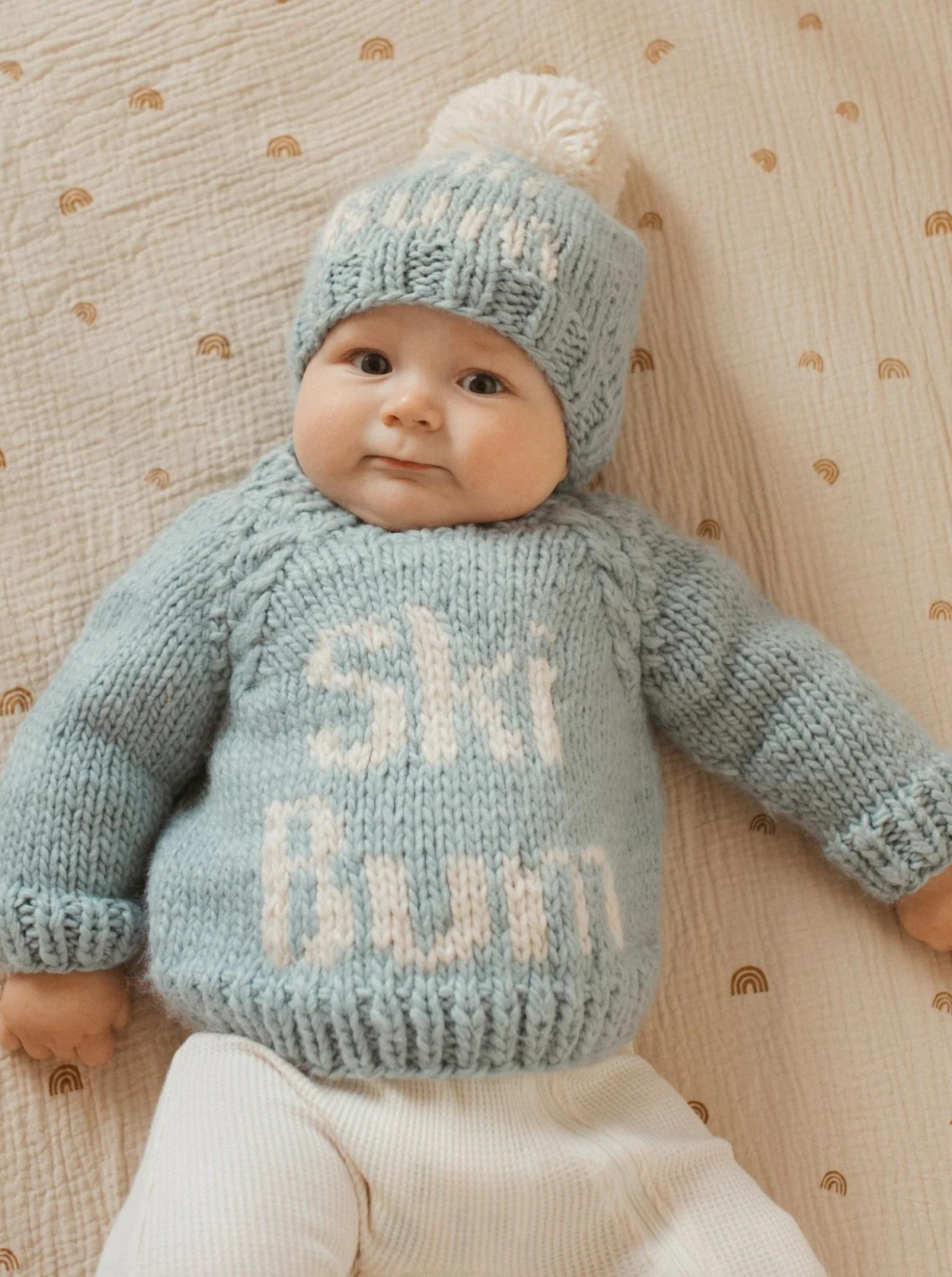 A cute baby wearing a light blue "Ski Bum" sweater and matching hat, resting on a patterned blanket.