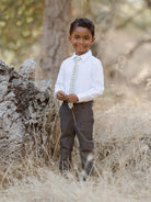 Smiling young boy in a white shirt and tie stands near a tree in a field of dry grass.