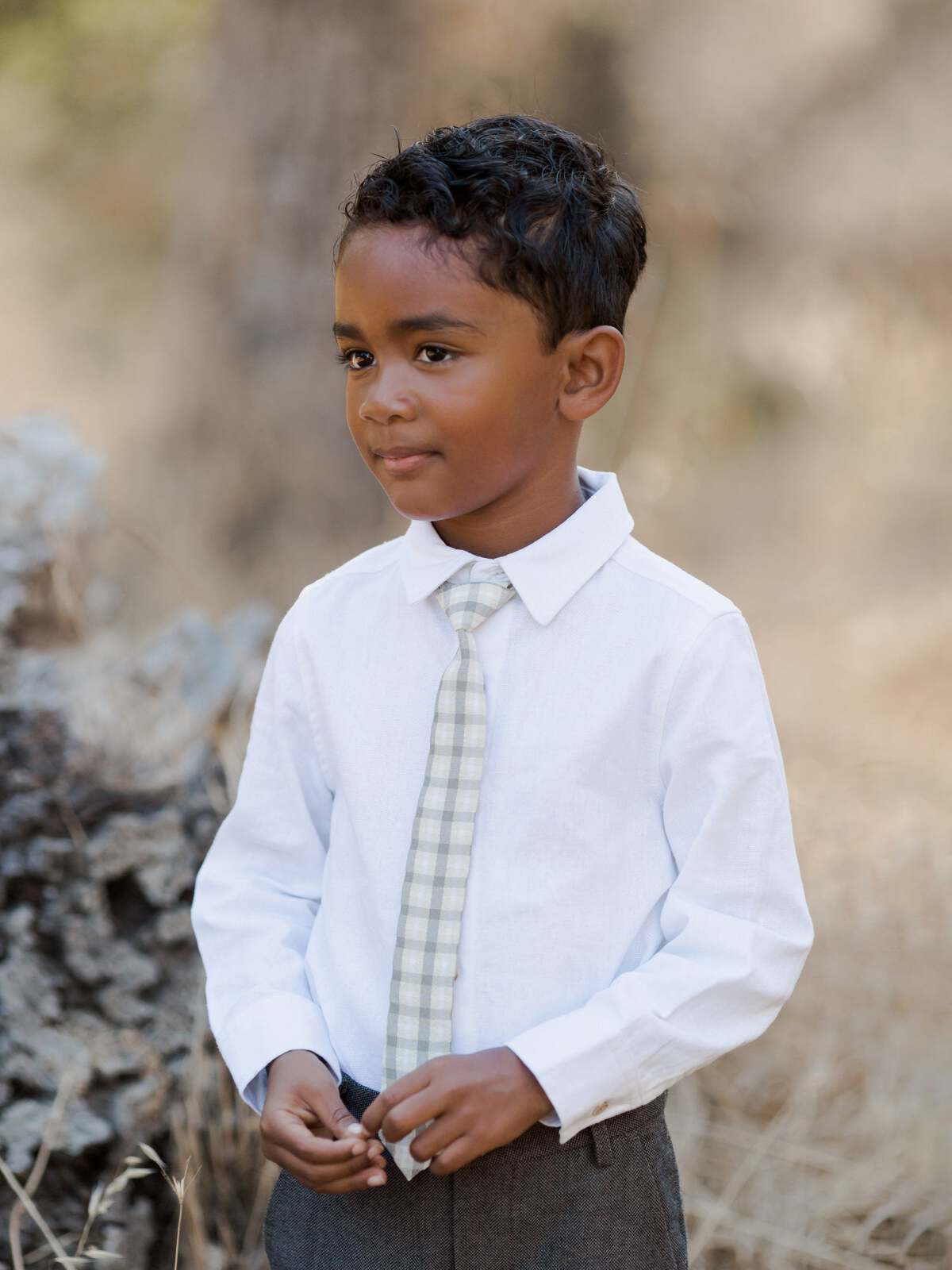 Young boy in a white shirt and plaid tie, standing outdoors with a neutral background.