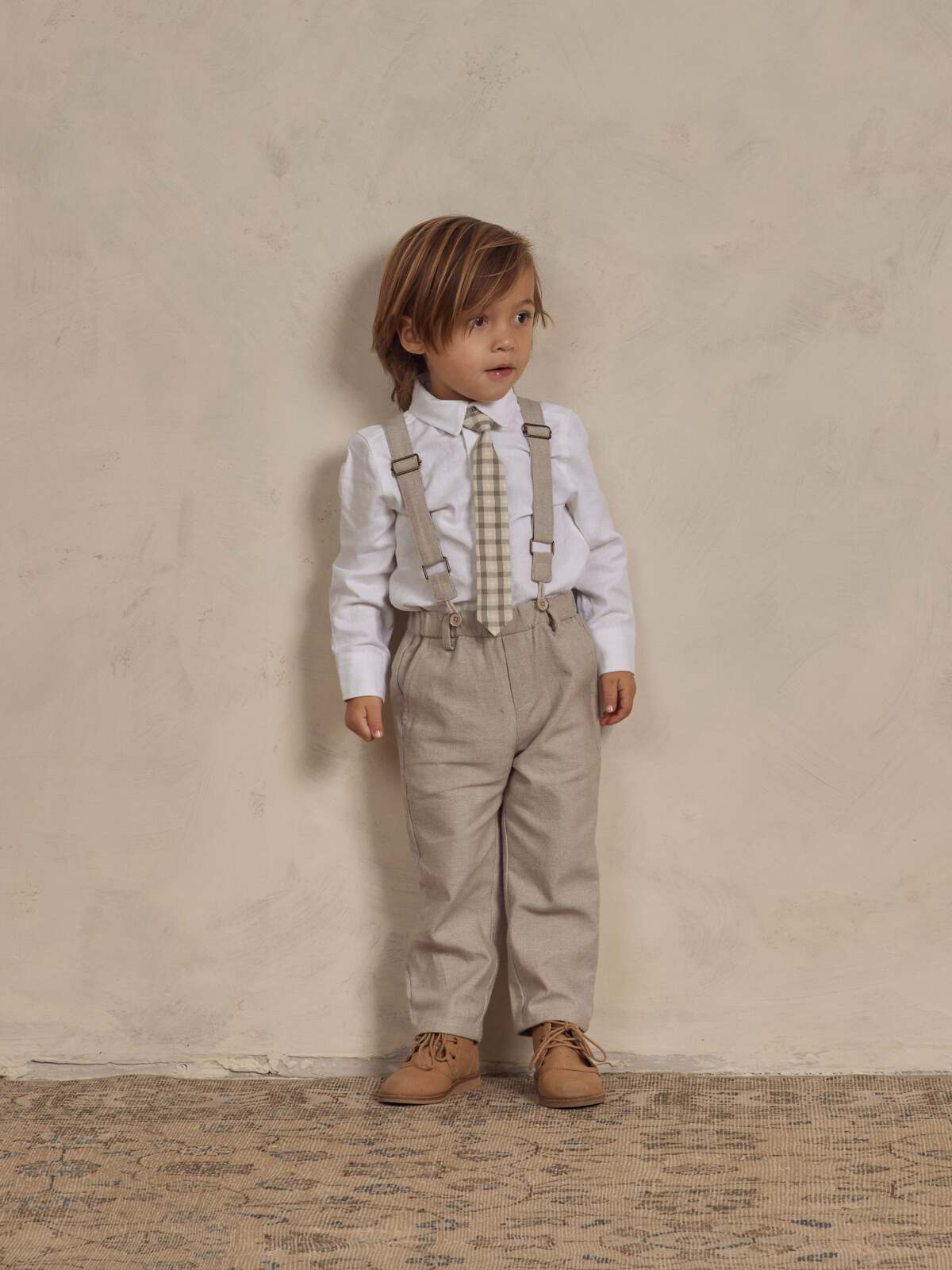 Child in beige pants, white shirt, and suspenders poses against a neutral background, showcasing a stylish outfit.