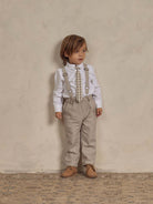 Child in beige pants, white shirt, and suspenders poses against a neutral background, showcasing a stylish outfit.