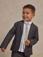 Young boy in a stylish gray suit and plaid tie, smiling against a soft beige background.