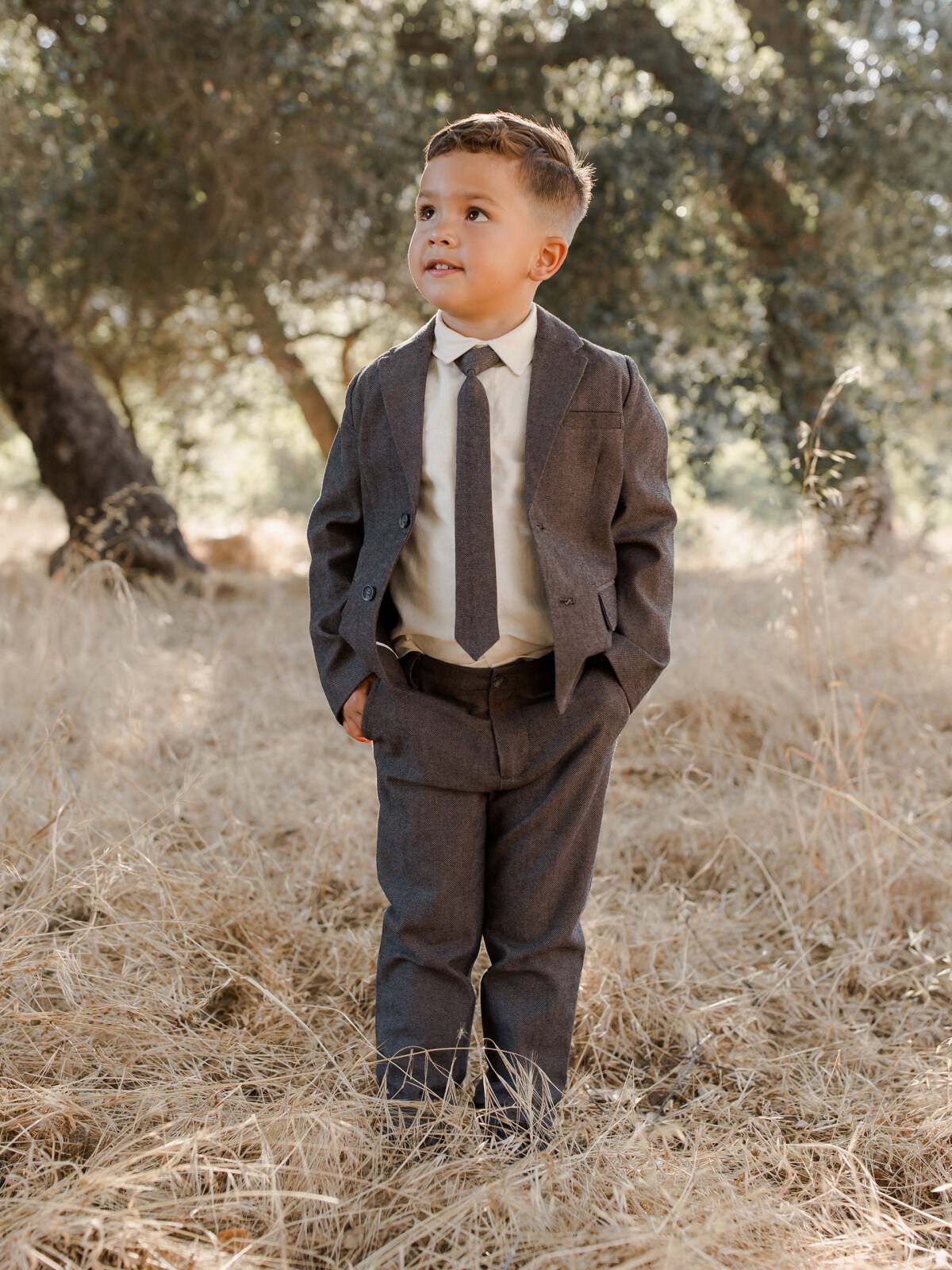 Boy in a suit stands confidently in a field of dry grass under trees, smiling and looking to the side.
