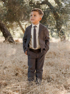 Boy in a suit stands confidently in a field of dry grass under trees, smiling and looking to the side.