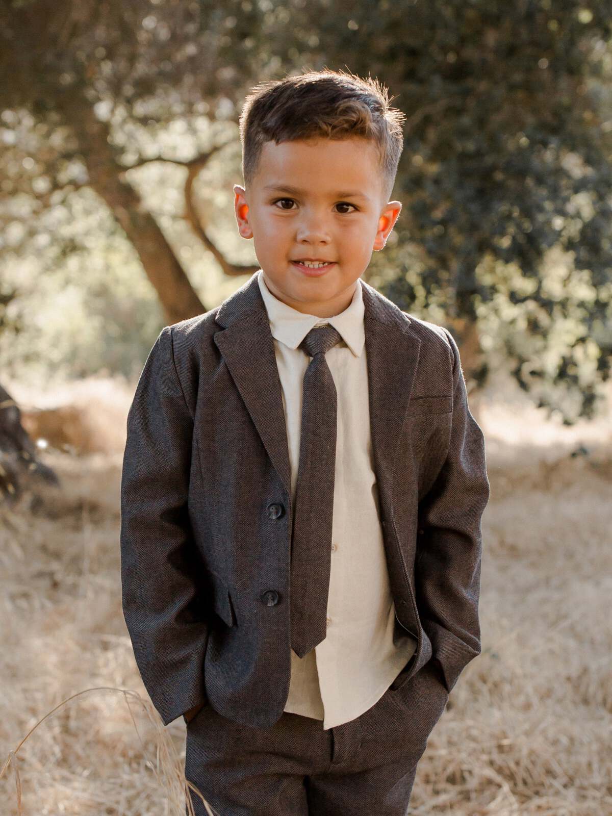 Young boy in a gray suit and tie, smiling outdoors in a grassy area with trees in the background.