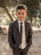 Young boy in a gray suit and tie, smiling outdoors in a grassy area with trees in the background.