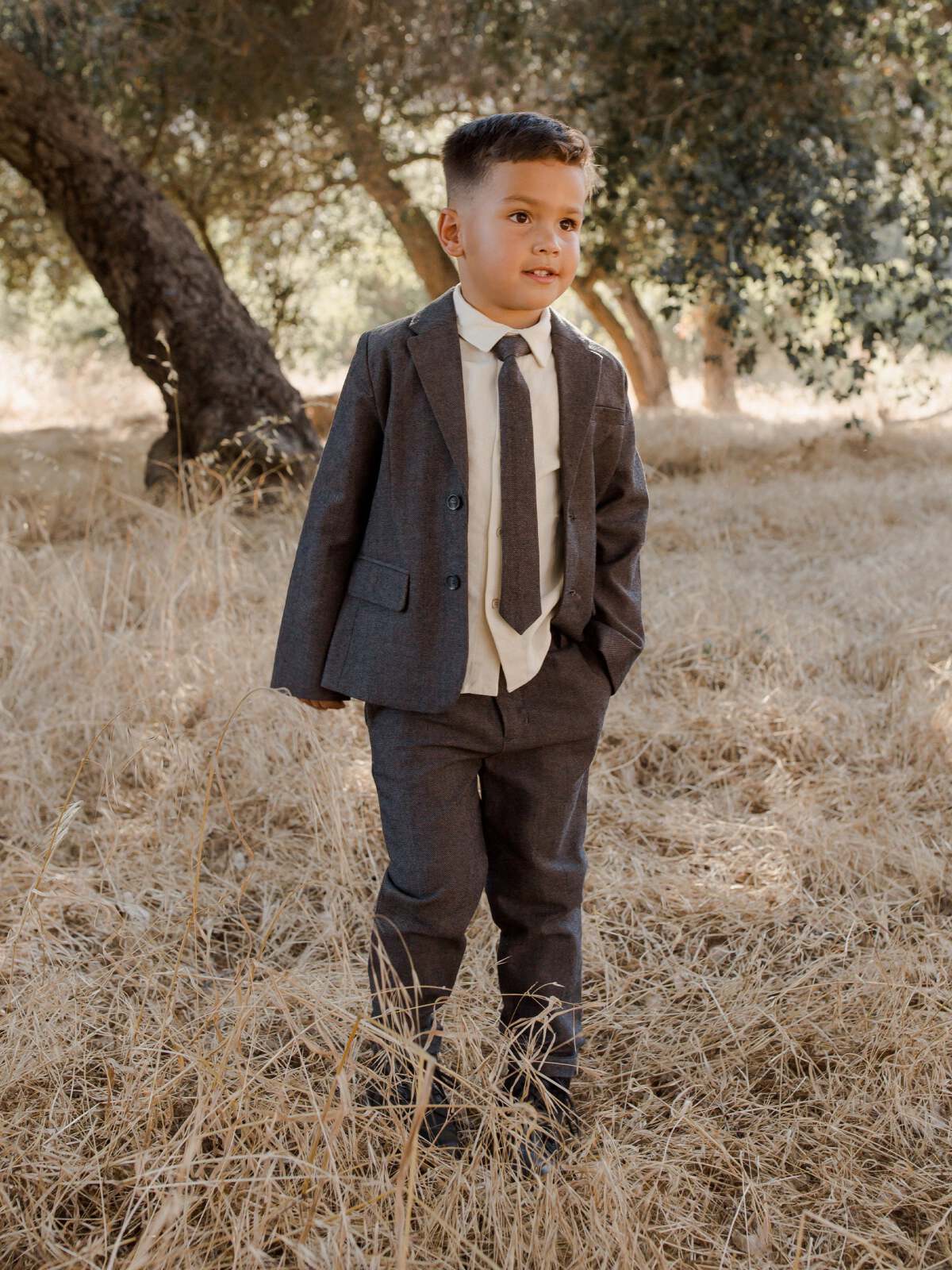 A young boy in a gray suit stands confidently in a field of tall grass, smiling in natural sunlight.