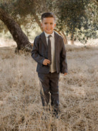 Smiling young boy in a suit standing in a grassy field, surrounded by trees.