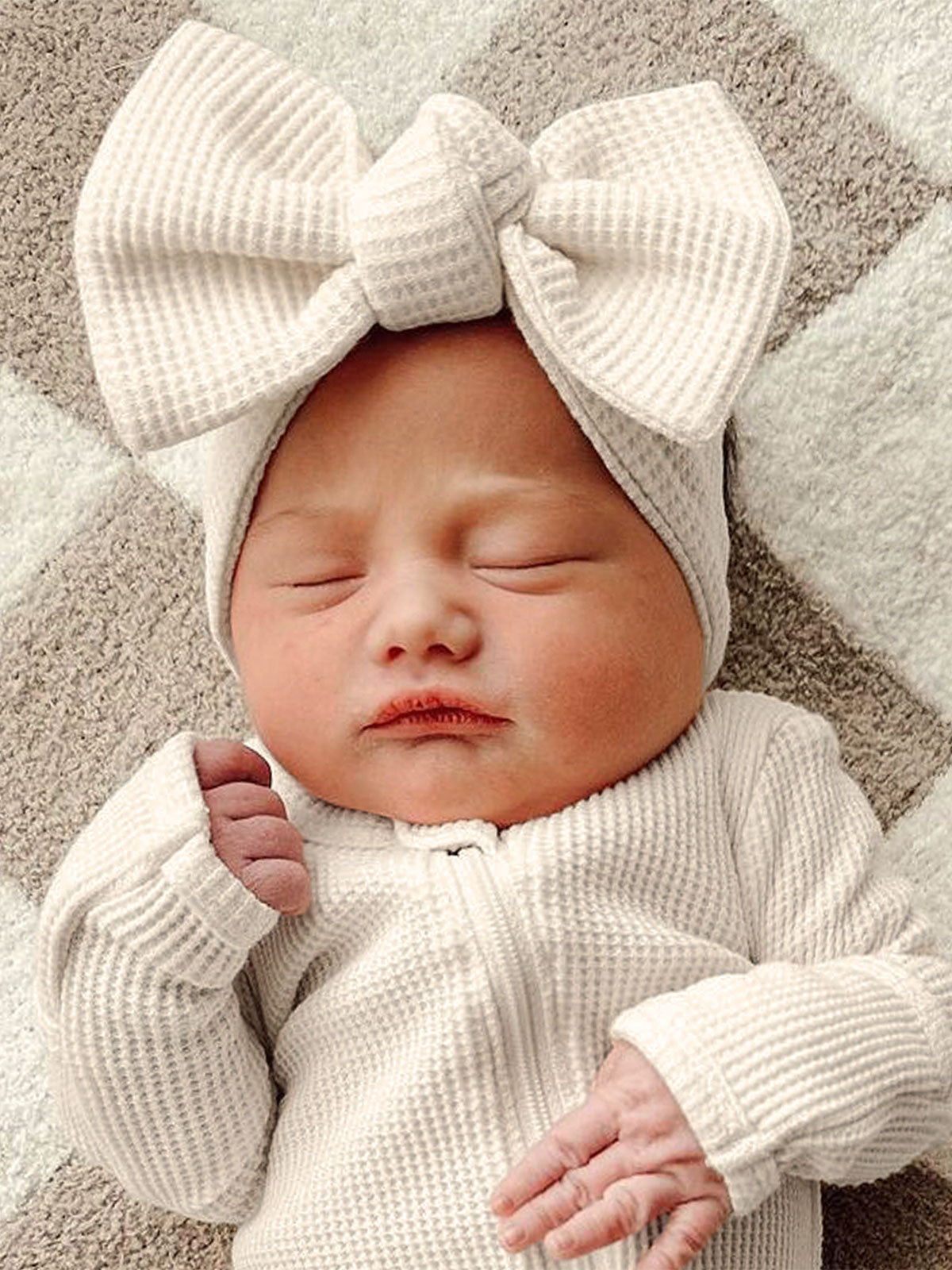 Newborn baby wearing a white outfit and large bow, peacefully sleeping on a soft, patterned blanket.