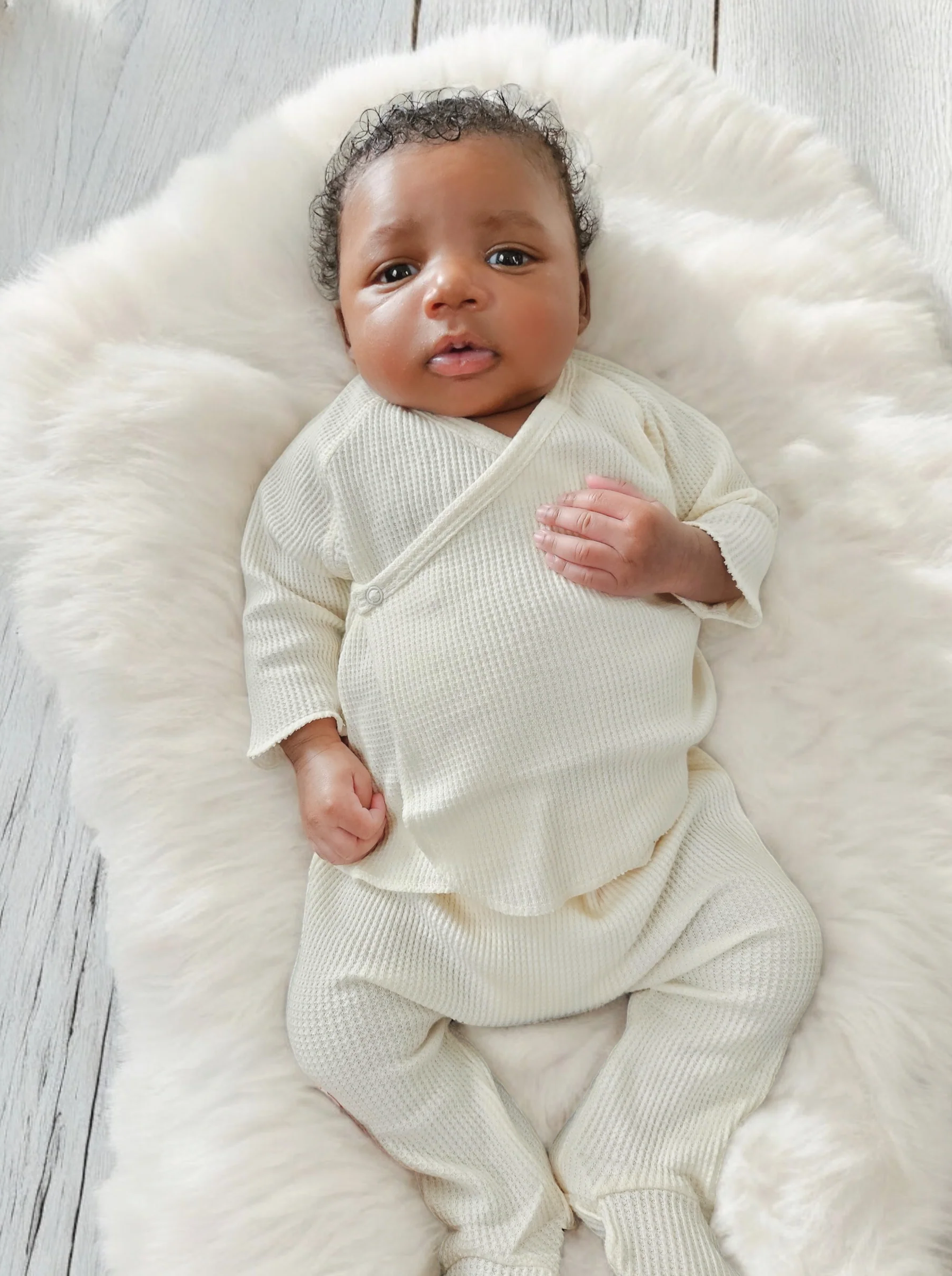 A newborn baby with curly hair, dressed in a cozy cream outfit, relaxing on a fluffy white blanket.