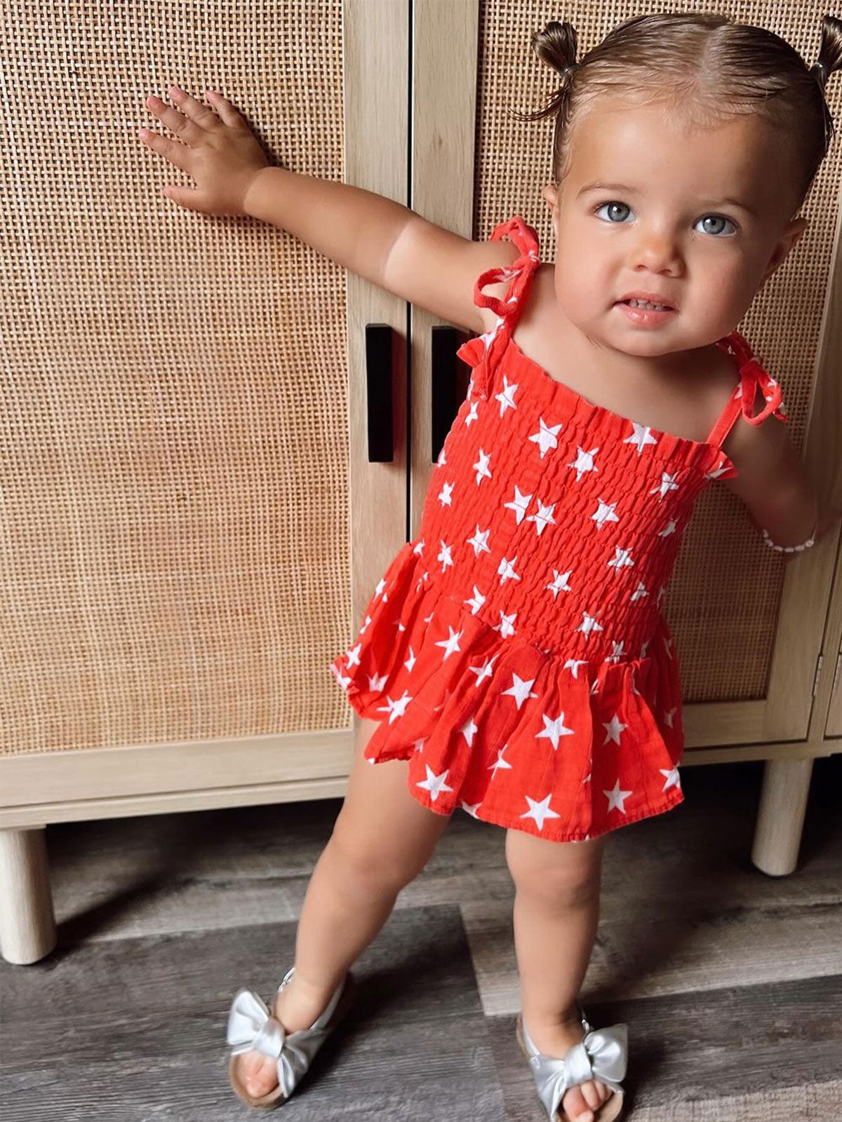Toddler in a red star-patterned outfit, standing by a wooden cabinet, smiling with pigtails and silver shoes.