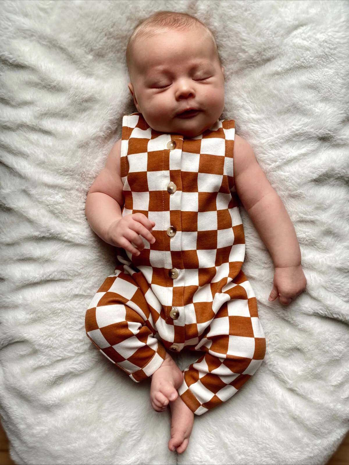 Baby sleeping on a soft blanket, wearing a brown and white checkered outfit, with a peaceful expression.
