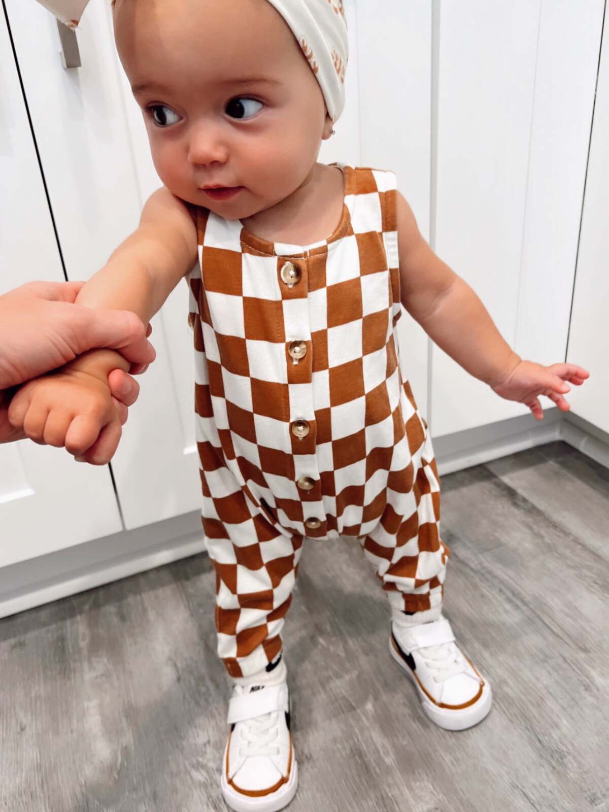 Toddler in a brown and white checked outfit, holding an adult's hand in a modern kitchen.