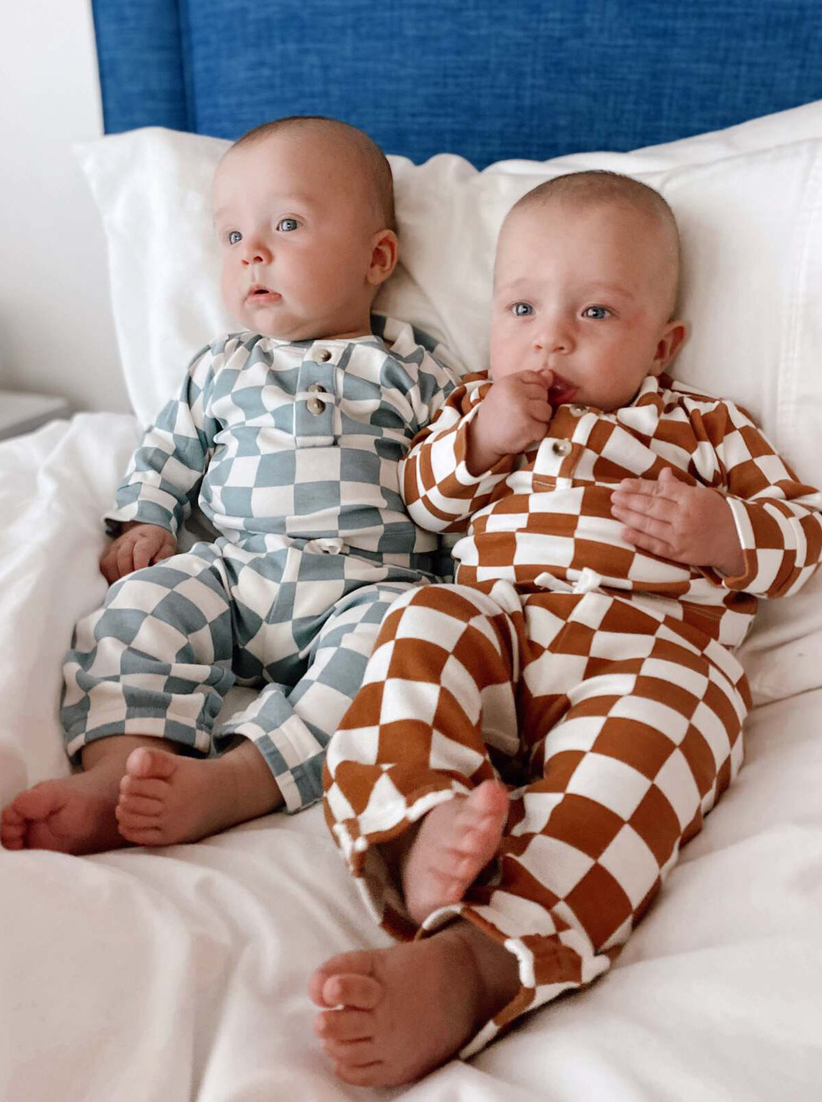 Two babies sitting on a bed, wearing patterned pajamas, with cute expressions and small feet visible.