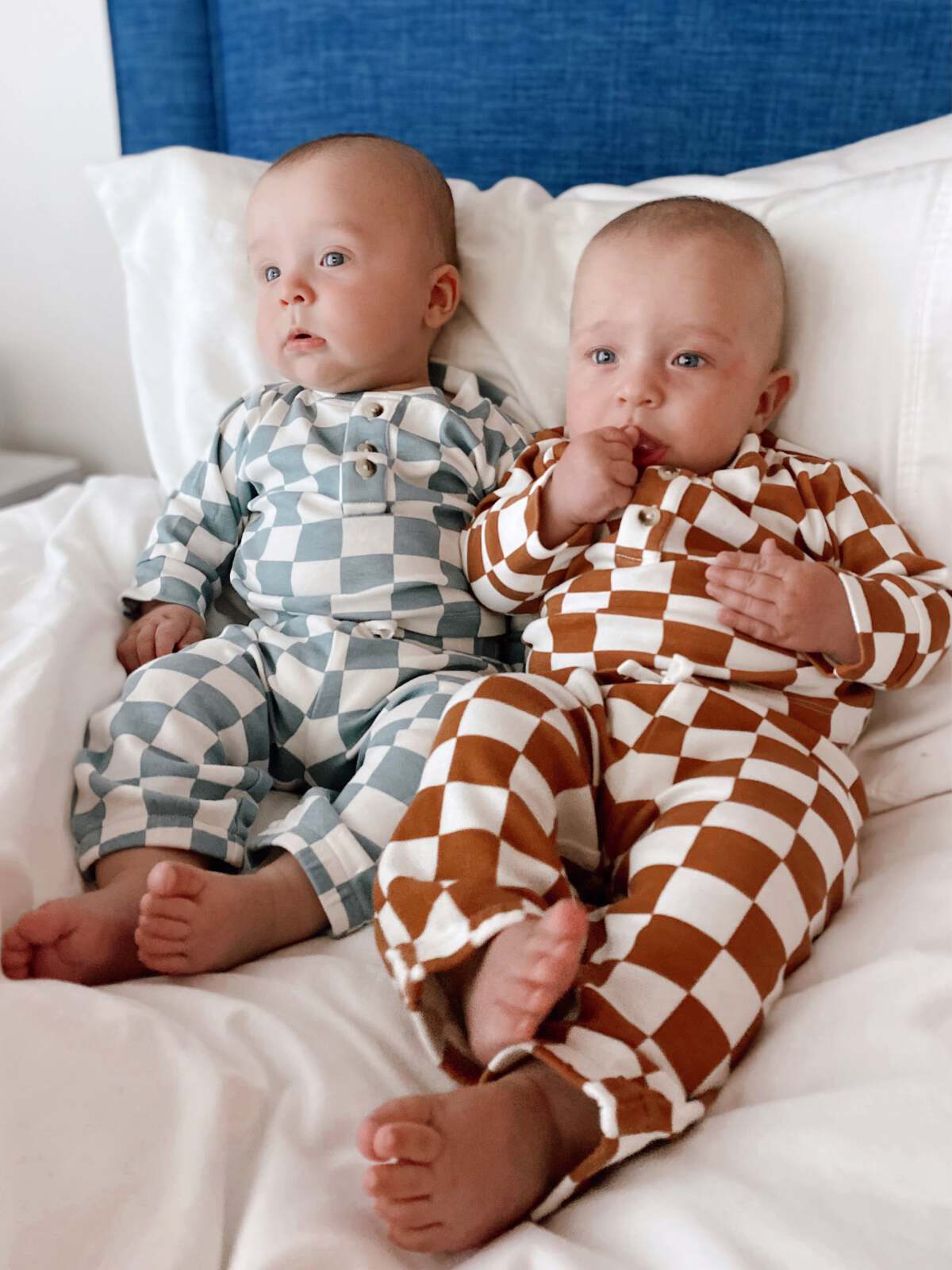 Two babies sitting on a bed, wearing patterned pajamas, with cute expressions and small feet visible.
