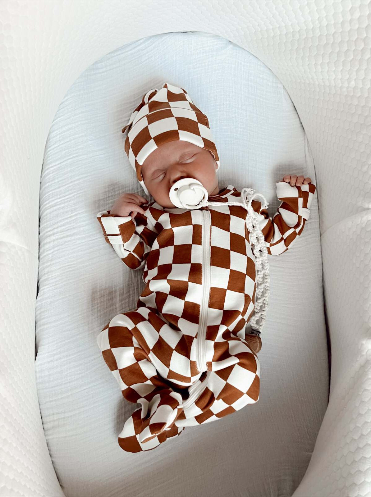 Baby peacefully sleeping in a brown and white checkered outfit with a matching hat, lying in a cozy bassinet.