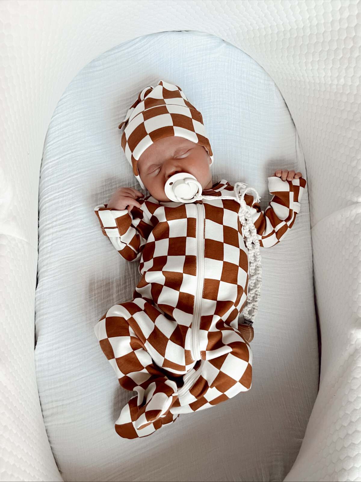 Baby peacefully sleeping in a brown and white checkered outfit with a matching hat, lying in a cozy bassinet.