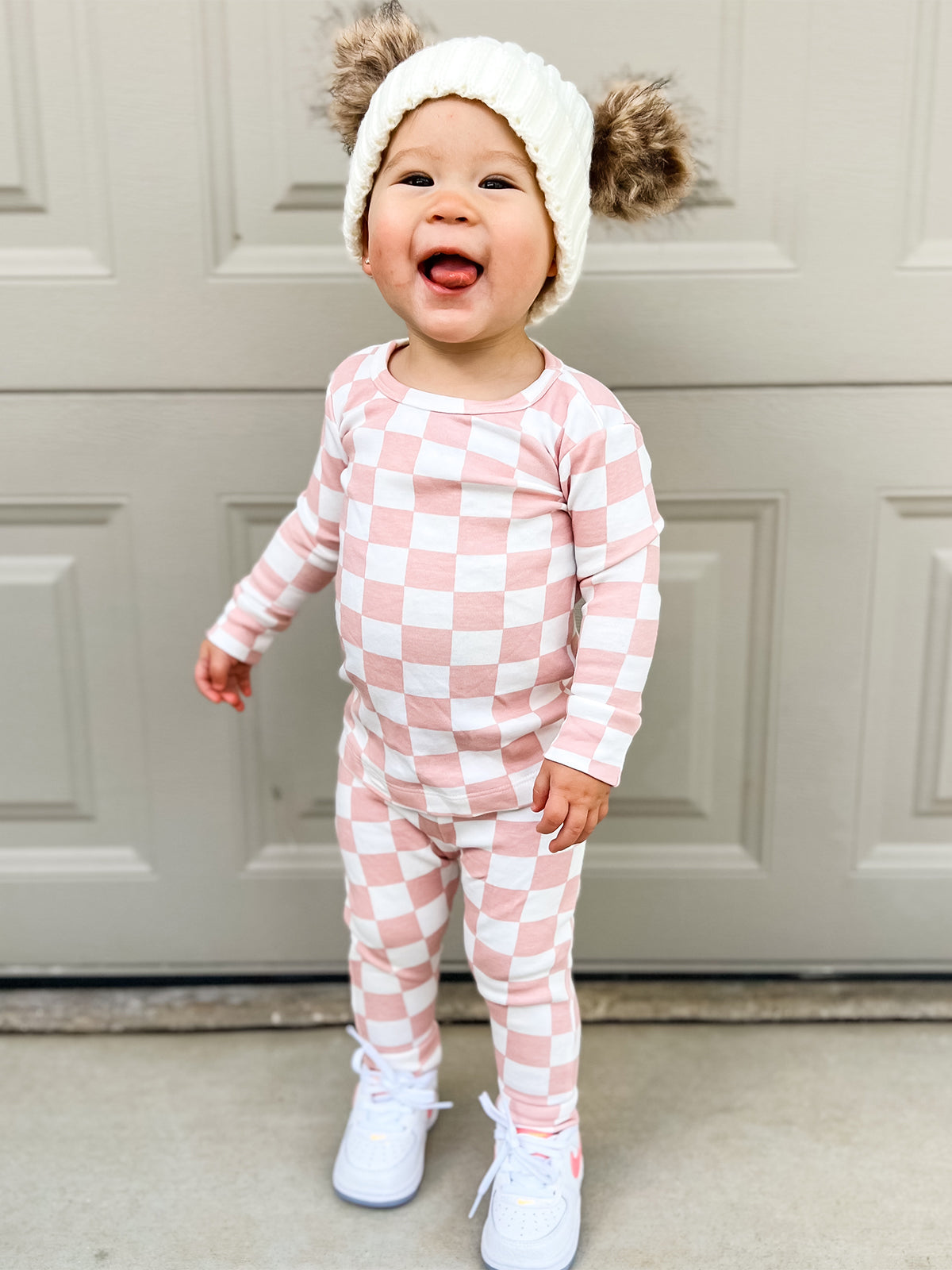 Smiling toddler in pink checkered outfit and fluffy hat stands on porch, wearing white sneakers.