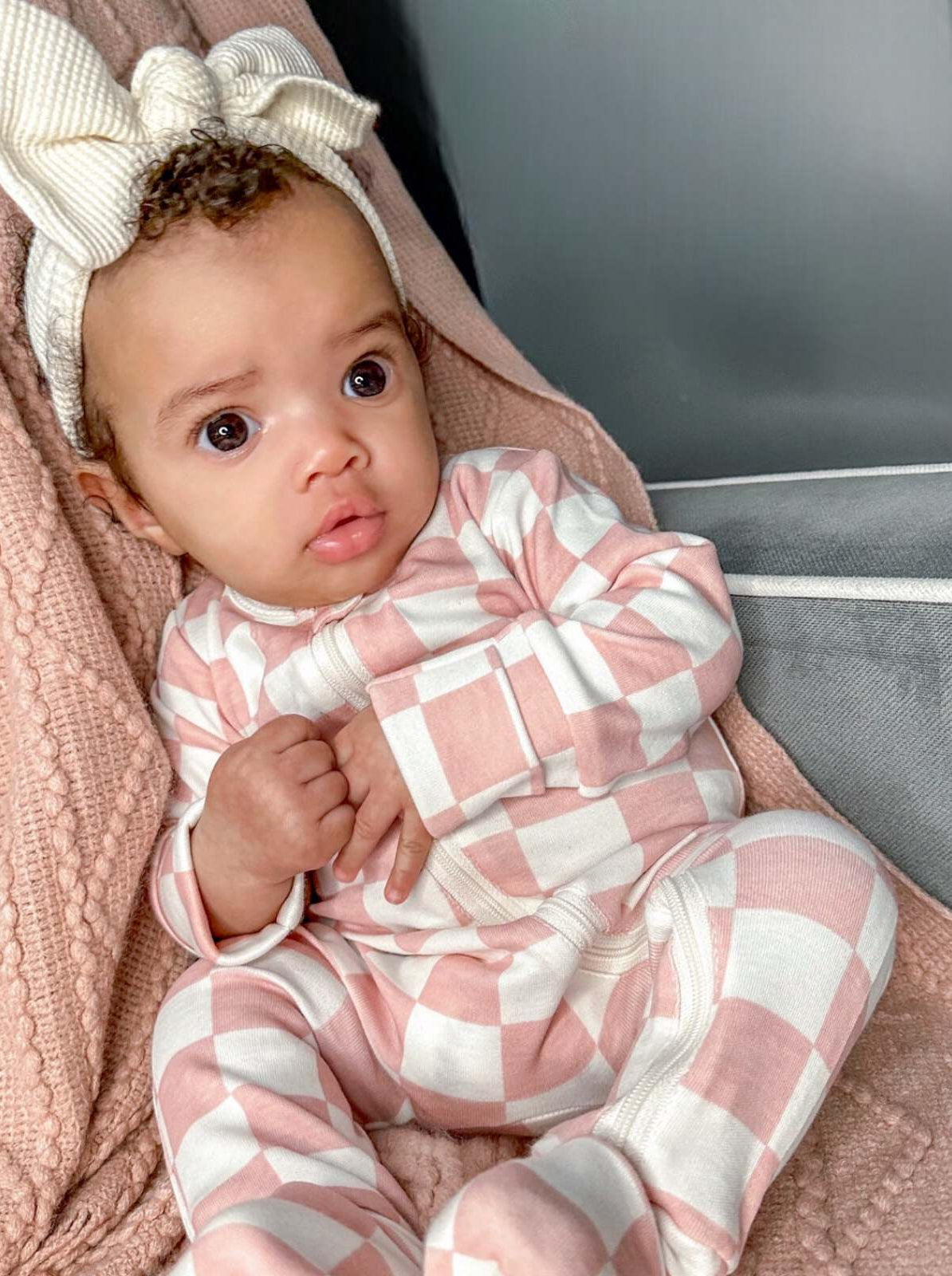 Baby in pink checkered pajamas with a large bow headband, sitting on a cozy blanket.