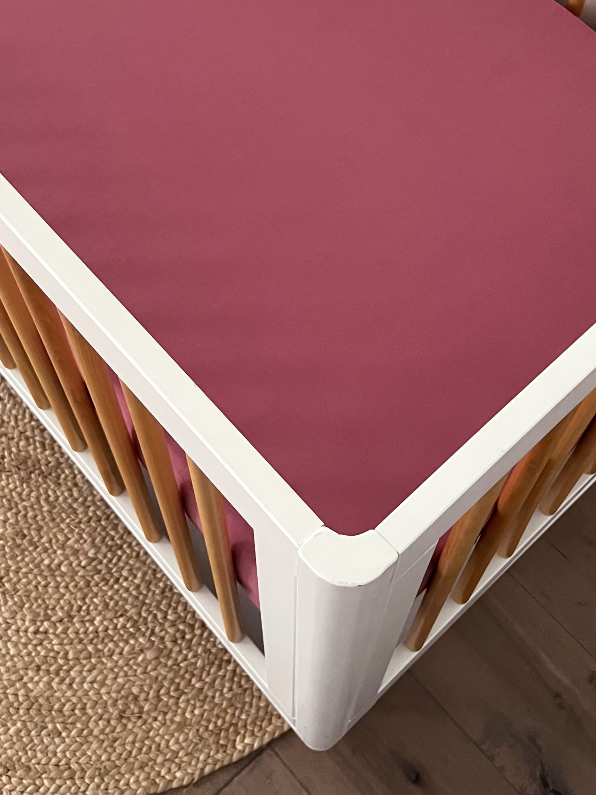 Top view of a modern crib with a burgundy mattress and light wood accents on a woven rug.