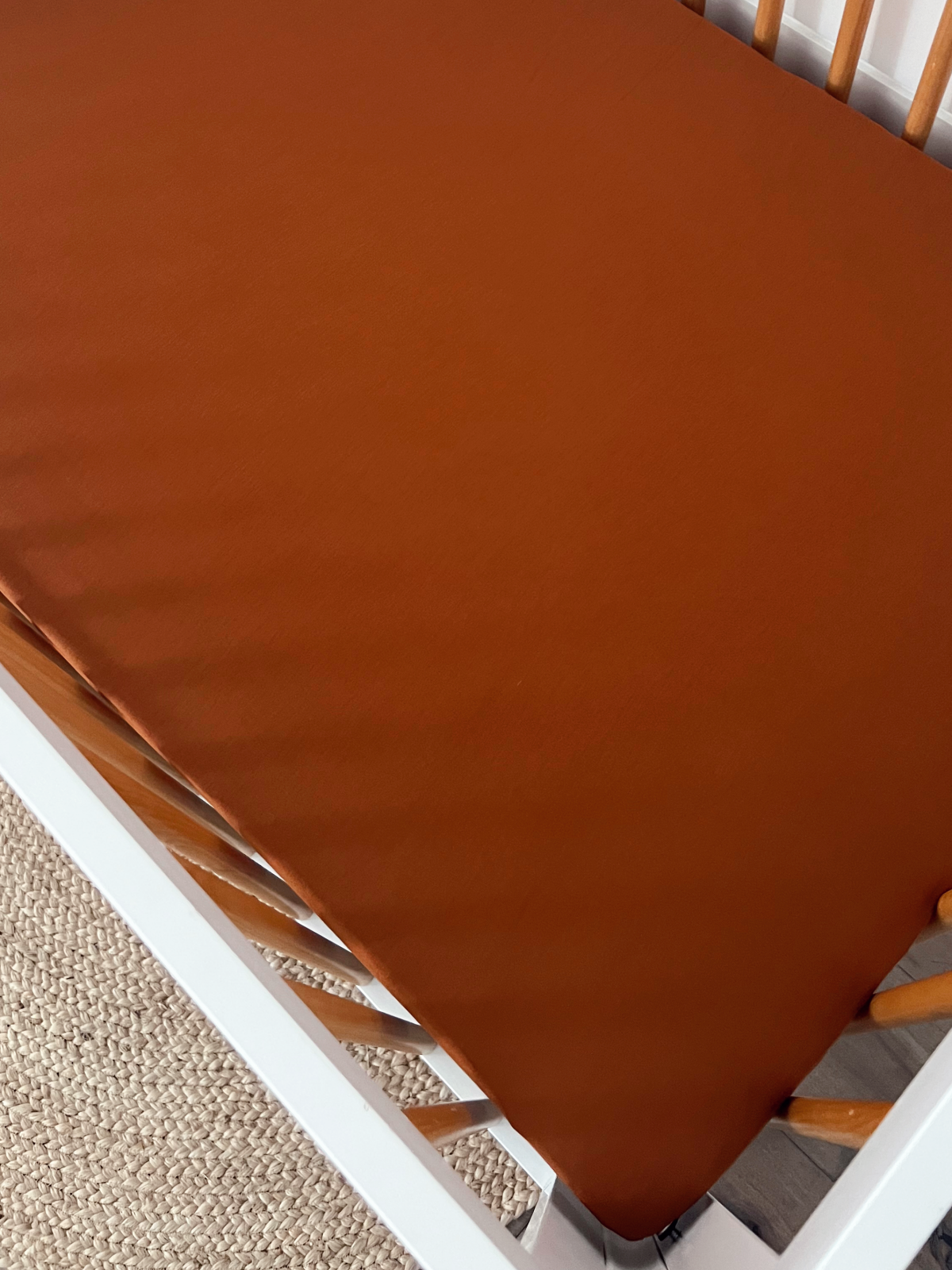 Rust-colored crib mattress cover in a wooden crib, with a woven rug visible underneath.