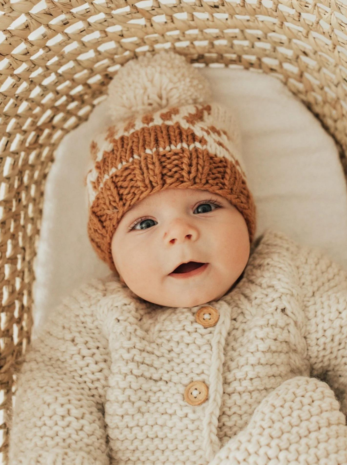 Smiling baby in cozy knitted sweater and hat, nestled in a woven basket.