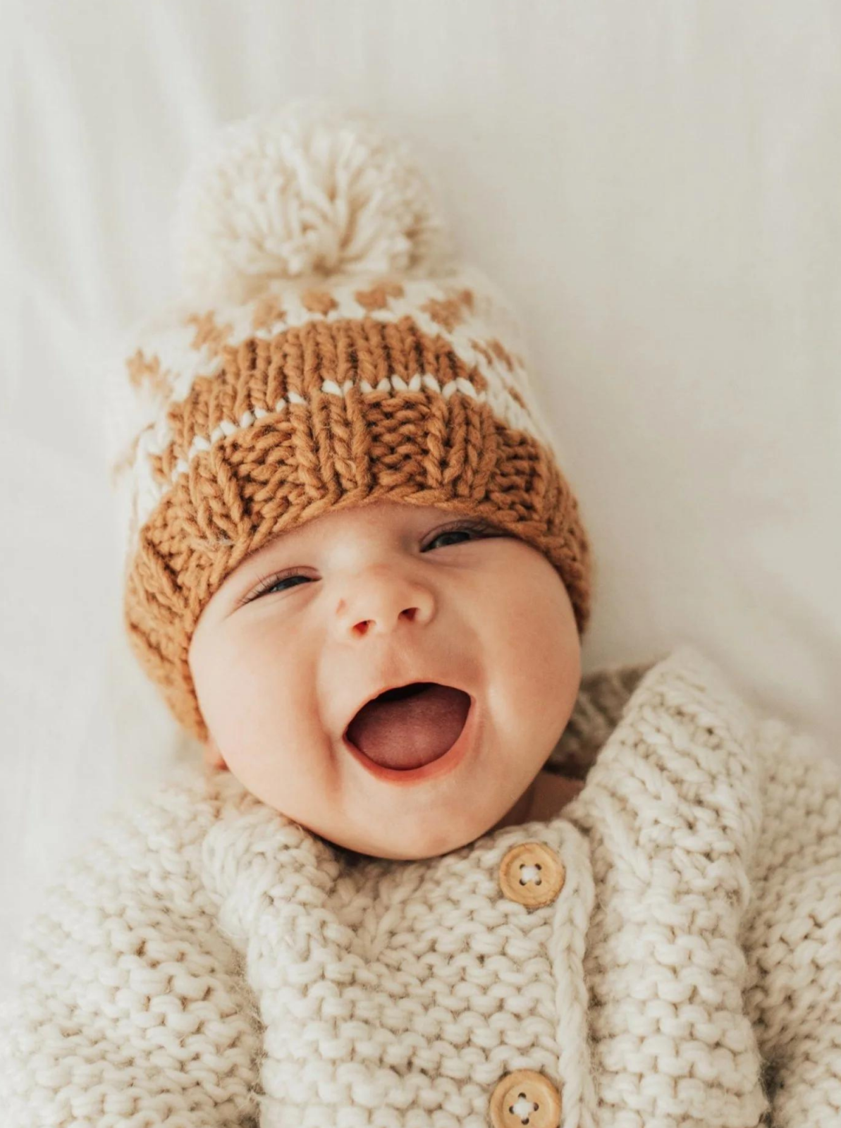 Smiling baby in a cozy knitted sweater and brown winter hat with a pom-pom, lying on a soft white surface.