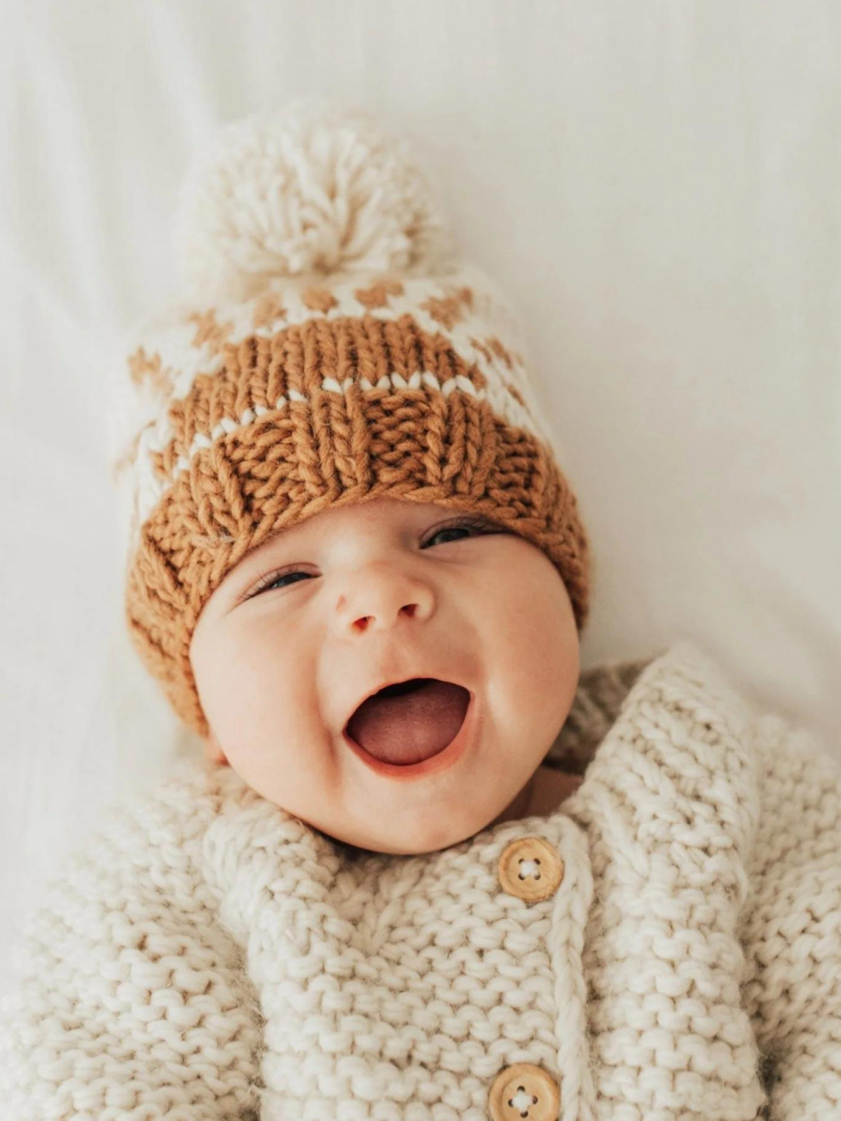 Smiling baby in a cozy knitted sweater and brown winter hat with a pom-pom, lying on a soft white surface.