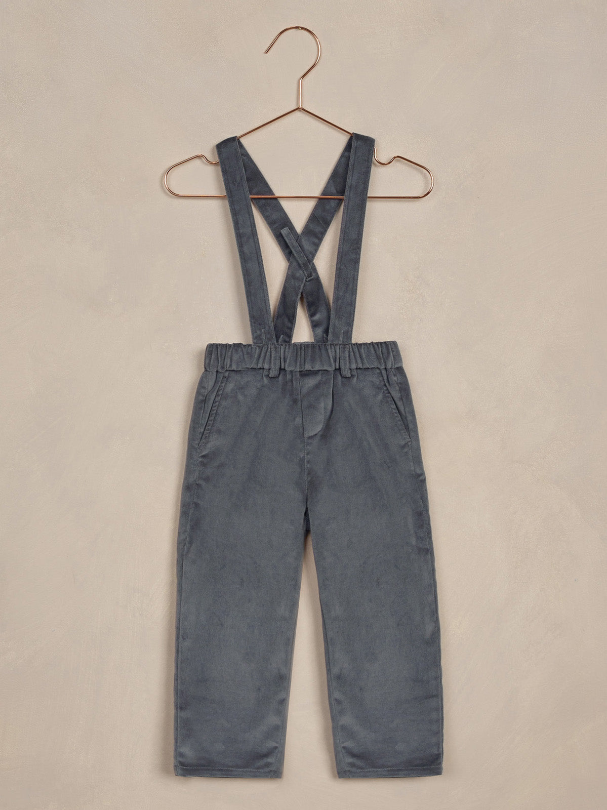 Gray overalls with crossed straps hanging on a copper hanger against a textured beige background.