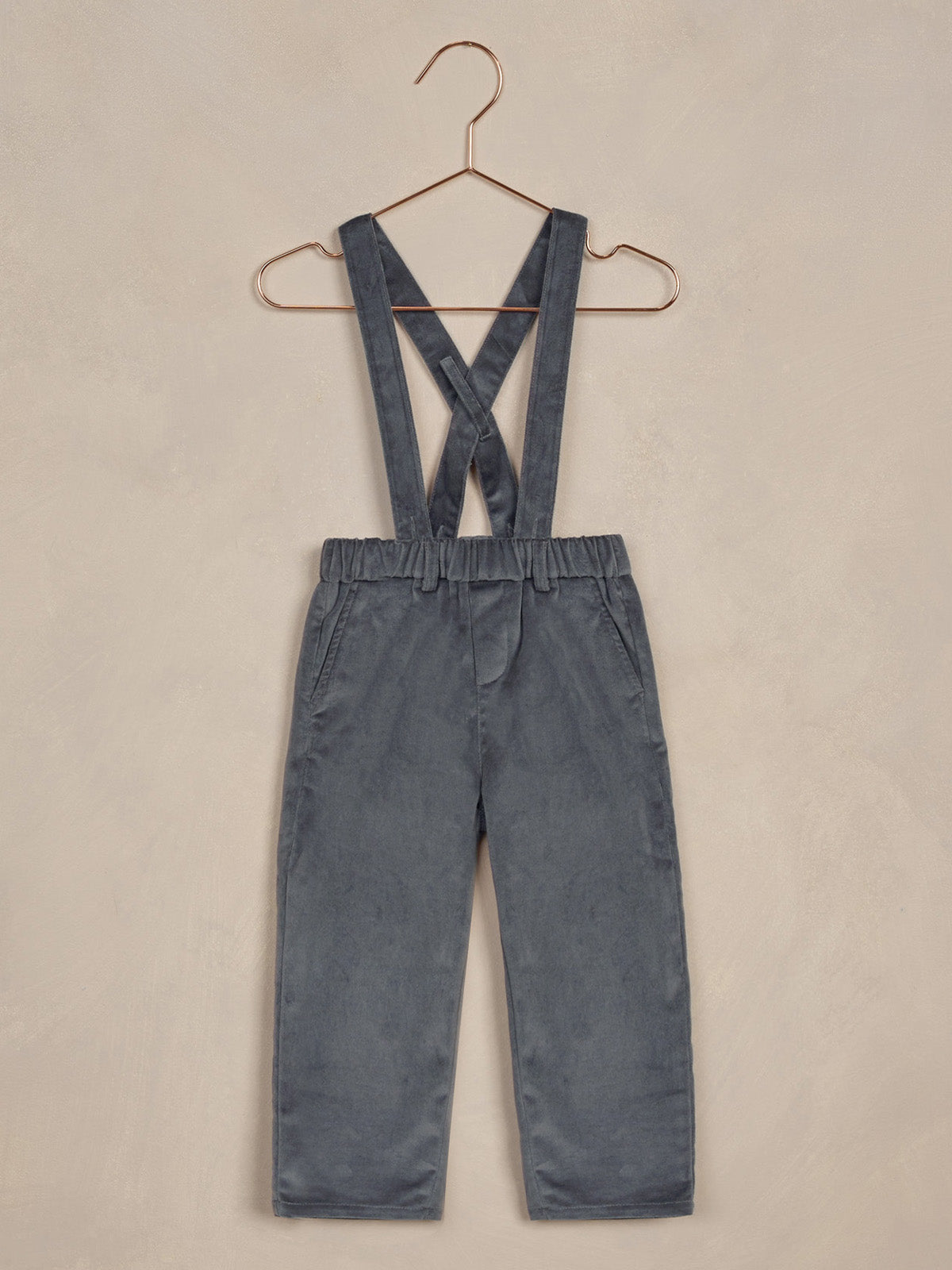 Gray overalls with crossed straps hanging on a copper hanger against a textured beige background.